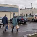 Zone commerciale près de Dieppe avec parking, façades sobres de magasins et un homme et une femme transportant leurs achats vers leurs voitures.